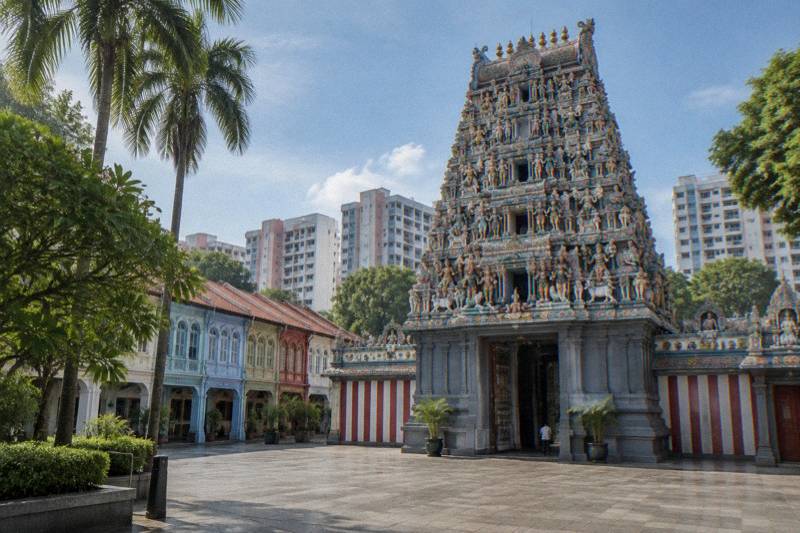 Morning at a Hindu temple in Chinatown, a tall pyramid-shaped gateway tower layered with colourful carved deity figures against a blue sky, a stone forecourt, one tiny distant figure.