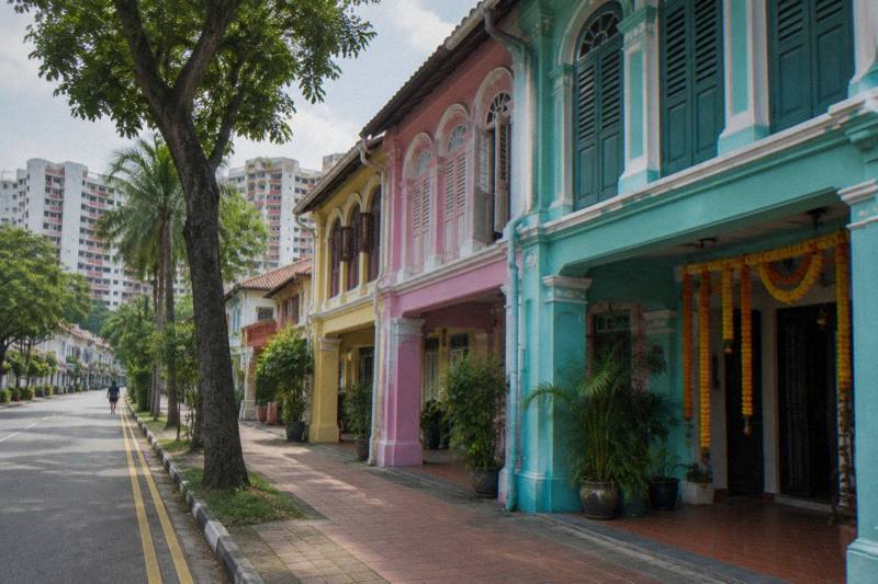 Morning on Serangoon Road in Little India, brightly coloured two-storey shophouses in turquoise, pink and yellow with arched windows, strings of marigold garlands hanging at a doorway, a clean pavement, one tiny distant figure.