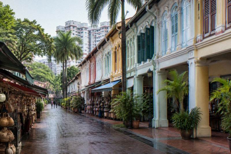 Bright morning on Pagoda Street in Chinatown, a row of pastel restored shophouses with shuttered windows and a covered walkway, market stalls under awnings along the pavement, damp ground, two tiny distant figures.