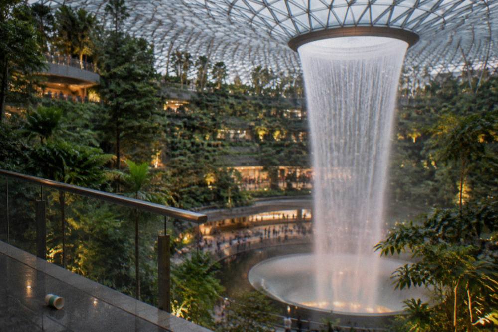 Shoppers and walkways circling the Rain Vortex waterfall inside Jewel Changi, with planted forest slopes on either side