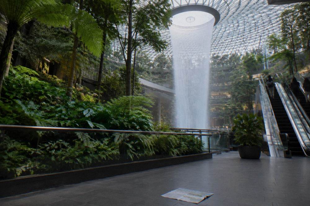 Planted garden slopes and walkways rising around the Rain Vortex inside Jewel Changi, seen from a lower public level