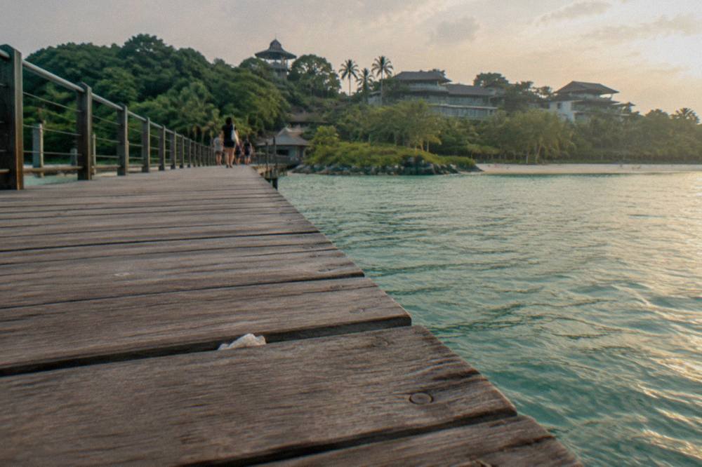 Wide wooden boardwalk crossing water toward a green island with resort buildings beyond