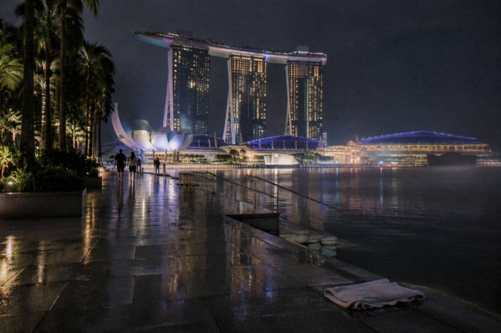 Open paved plaza beside a bay with lit hotel towers rising above the water at night