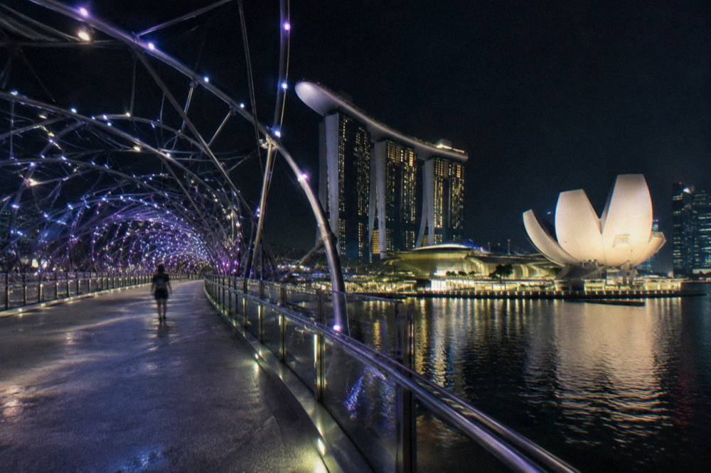 Curved pedestrian bridge lit at night above a bay with city towers glowing across the water