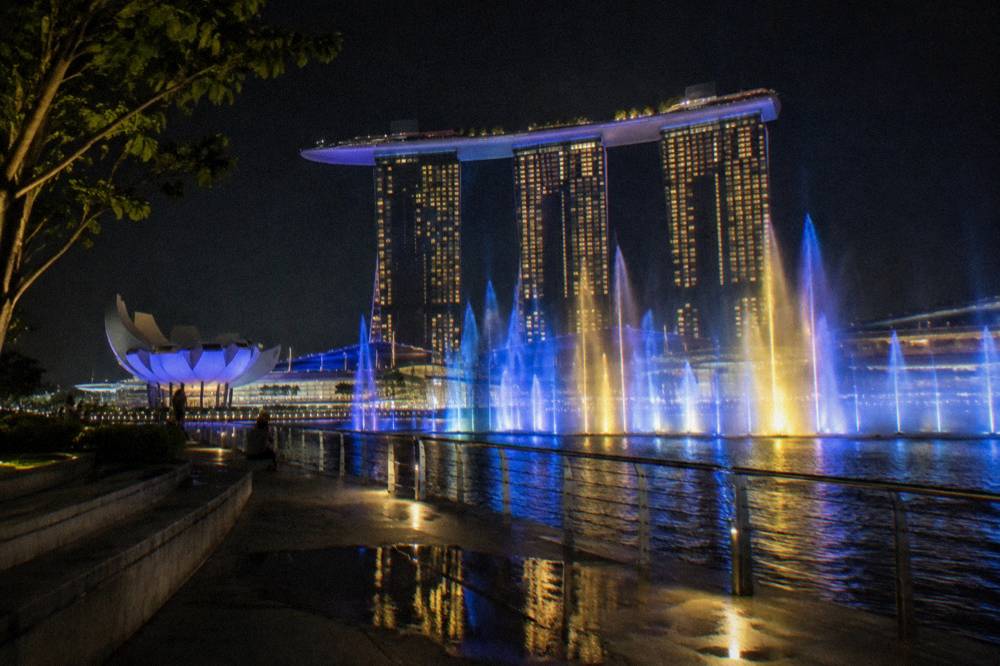 Water jets and coloured lights rising over a dark bay in front of lit towers