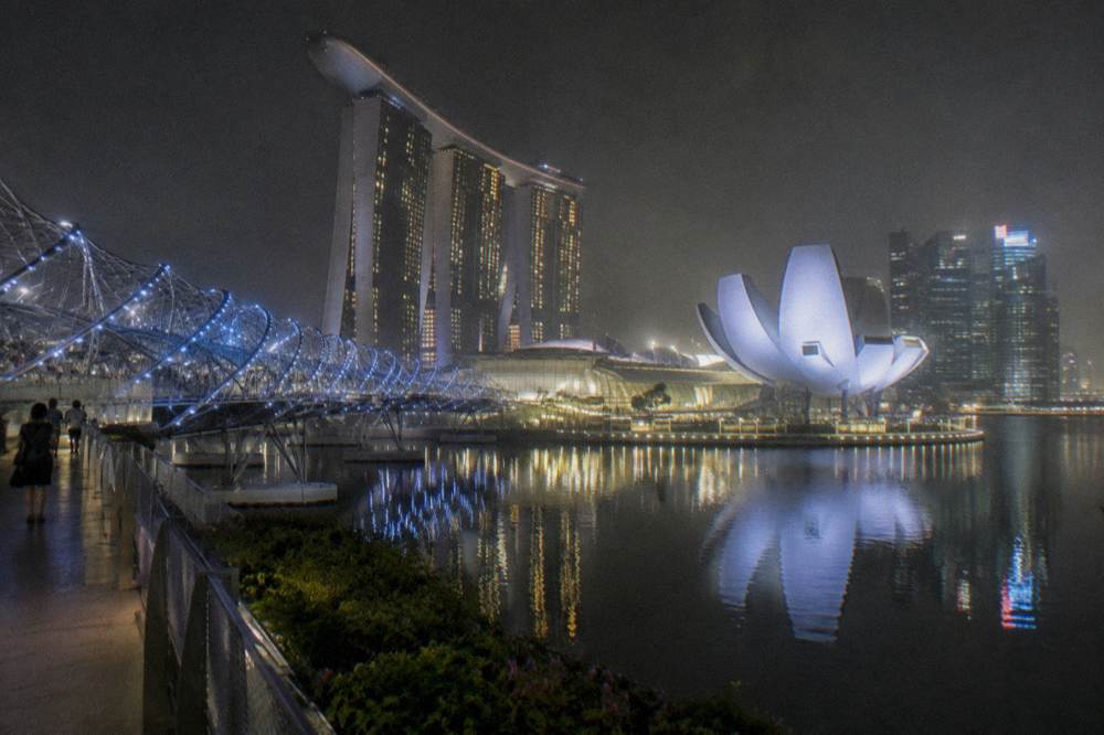 A lit curved bridge and illuminated towers reflected in still bay water at night